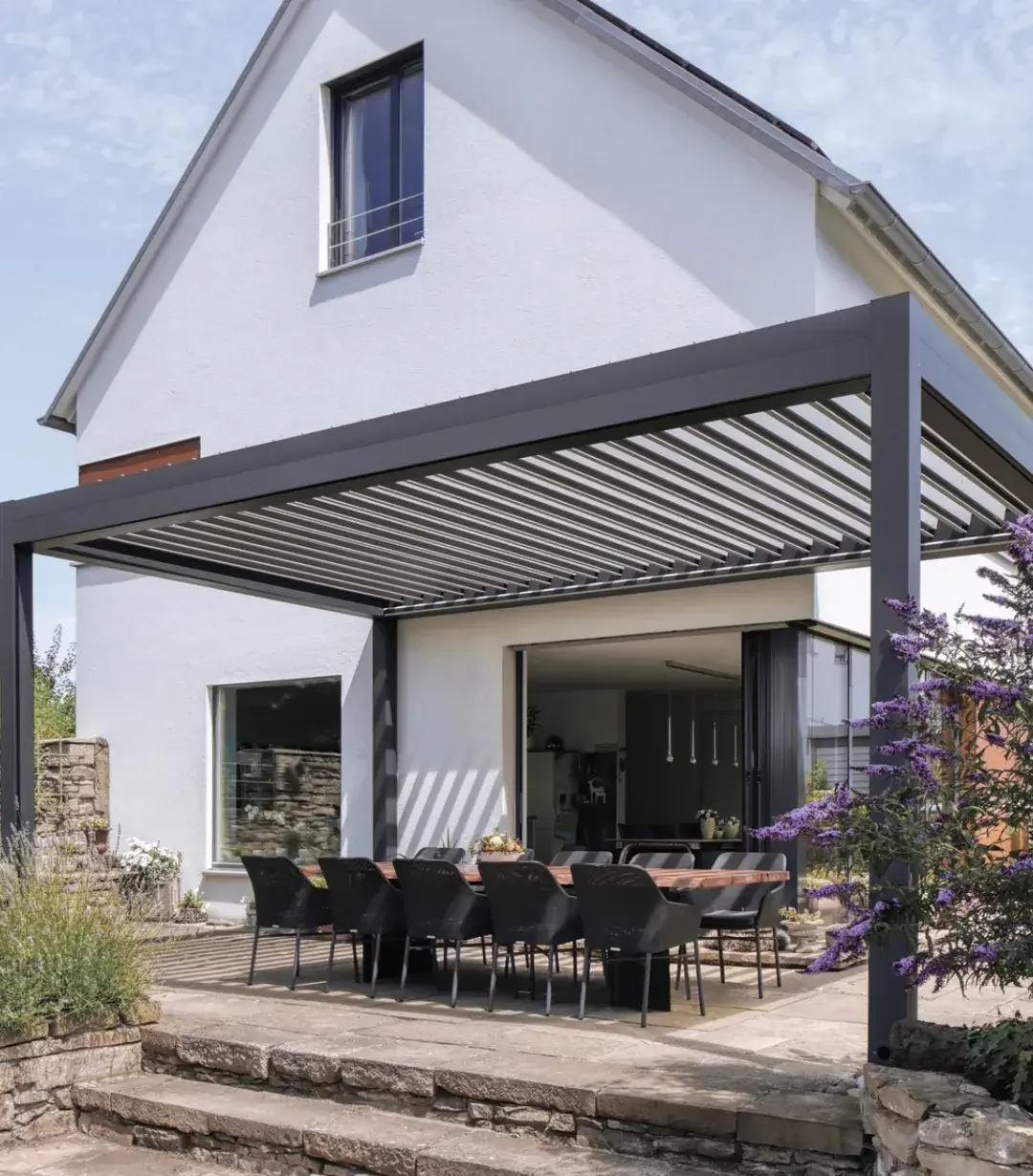 Lamellendach in Anthrazit mit verstellbaren Aluminiumlamellen an einem weißen Haus, darunter ein langer Esstisch mit schwarzen Stühlen